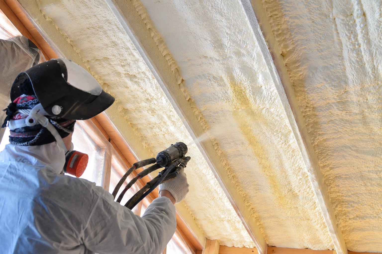 A worker in protective gear sprays foam onto the underside of a roof, filling the spaces between rafters.