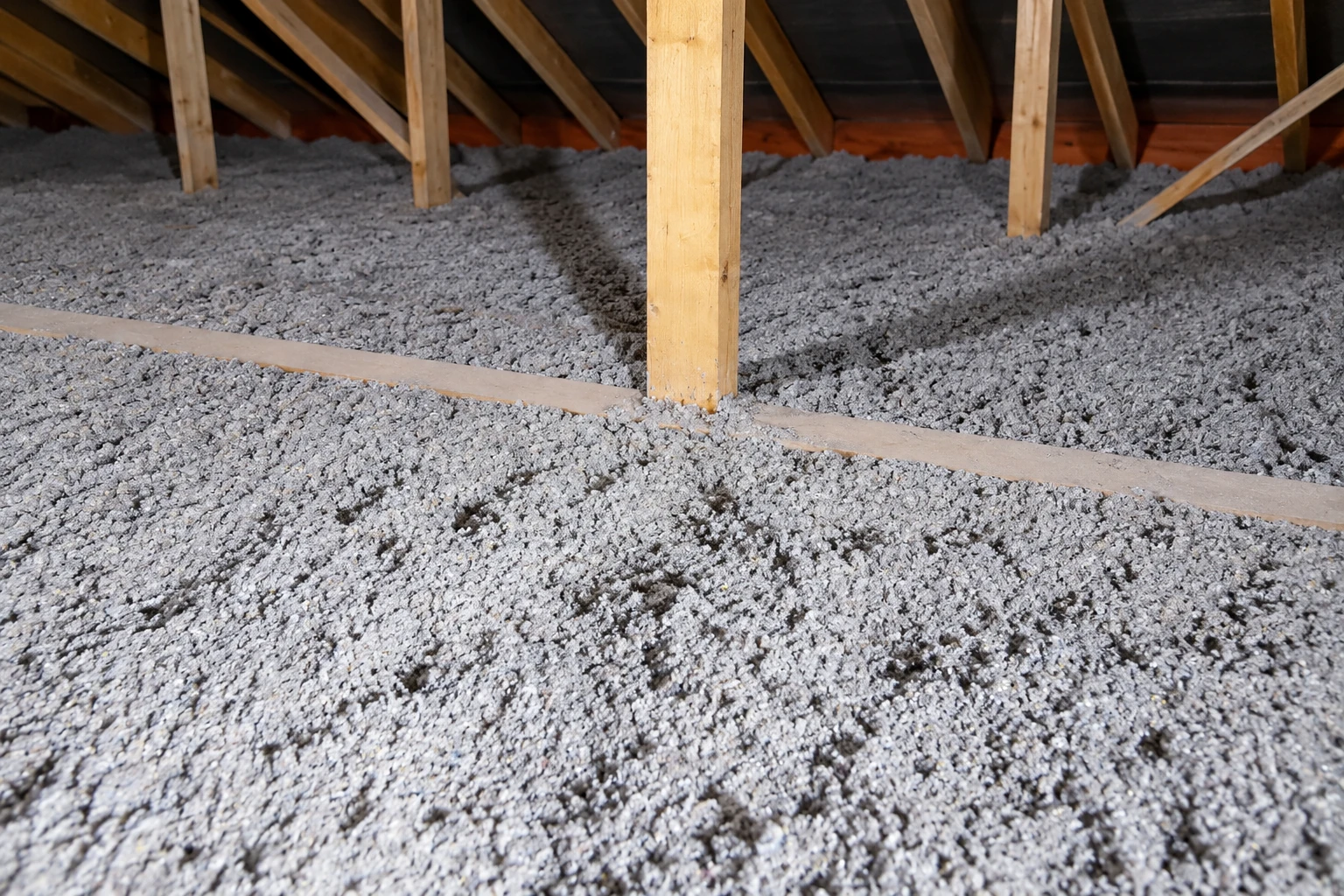 Loose gray insulation covers an attic floor beneath exposed wooden roof supports.