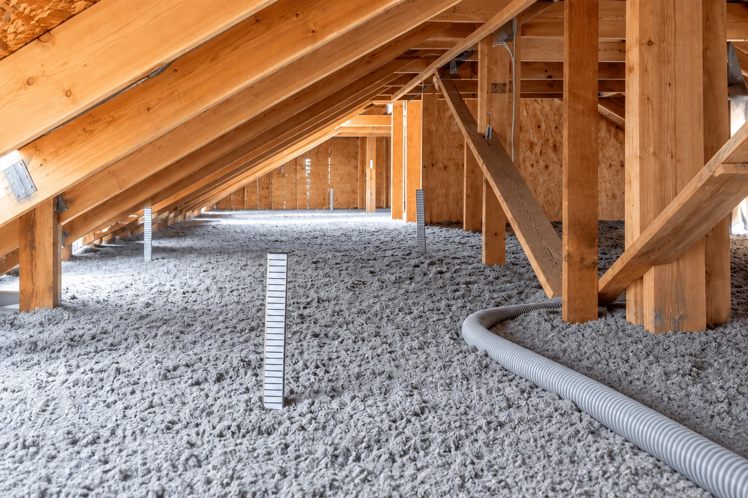 Blown-in insulation covers an attic floor, with a flexible hose and upright depth markers among the rafters.