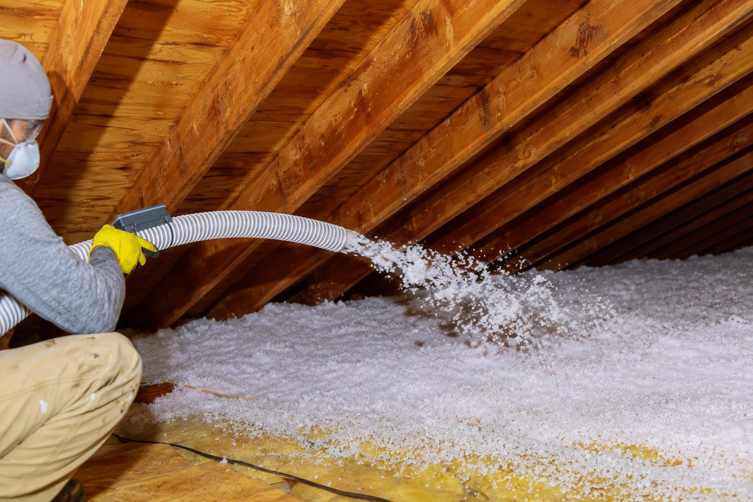 Worker directs a hose to blow loose-fill insulation across an attic floor beneath exposed wooden rafters.