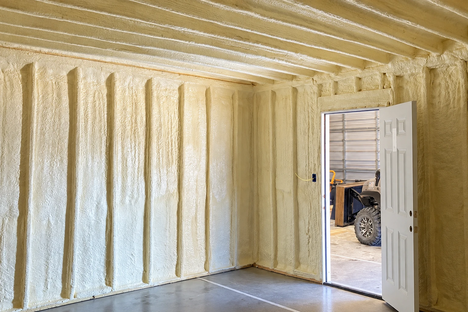 A commercial space has pale spray foam insulation covering its walls and ceiling, with an open door at right.