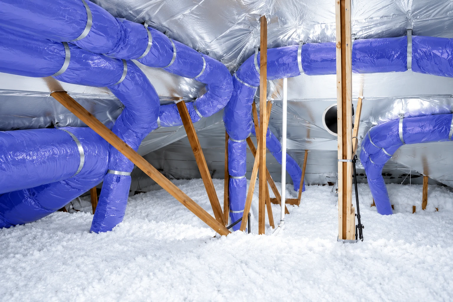 Blue flexible ducts run through a foil-lined attic above a thick layer of loose white insulation.