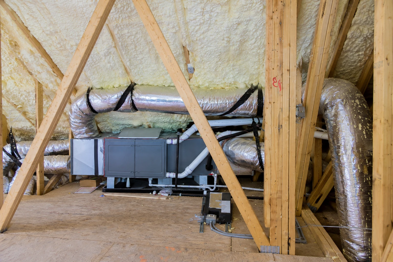 Attic HVAC unit and silver ductwork sit among wooden roof trusses, with spray foam covering the roof underside.