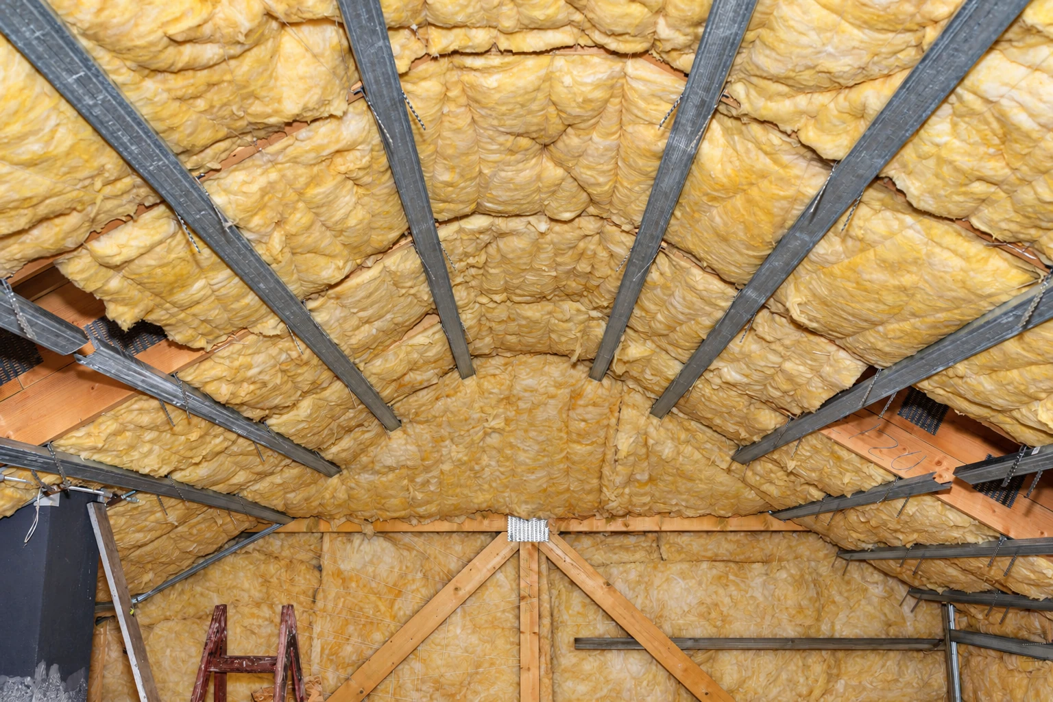 Yellow fiberglass batts fill the rafters of a roof, held between gray metal framing and exposed wood trusses.