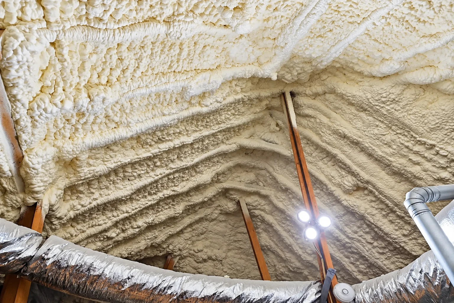 Attic roof and rafters covered in thick, uneven white spray foam, with metal ductwork and a work light below.